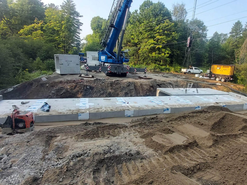 Une grue est utilisée pour installer des dalles de béton préfabriquées sur un chantier de construction de pont, entouré de véhicules de chantier.